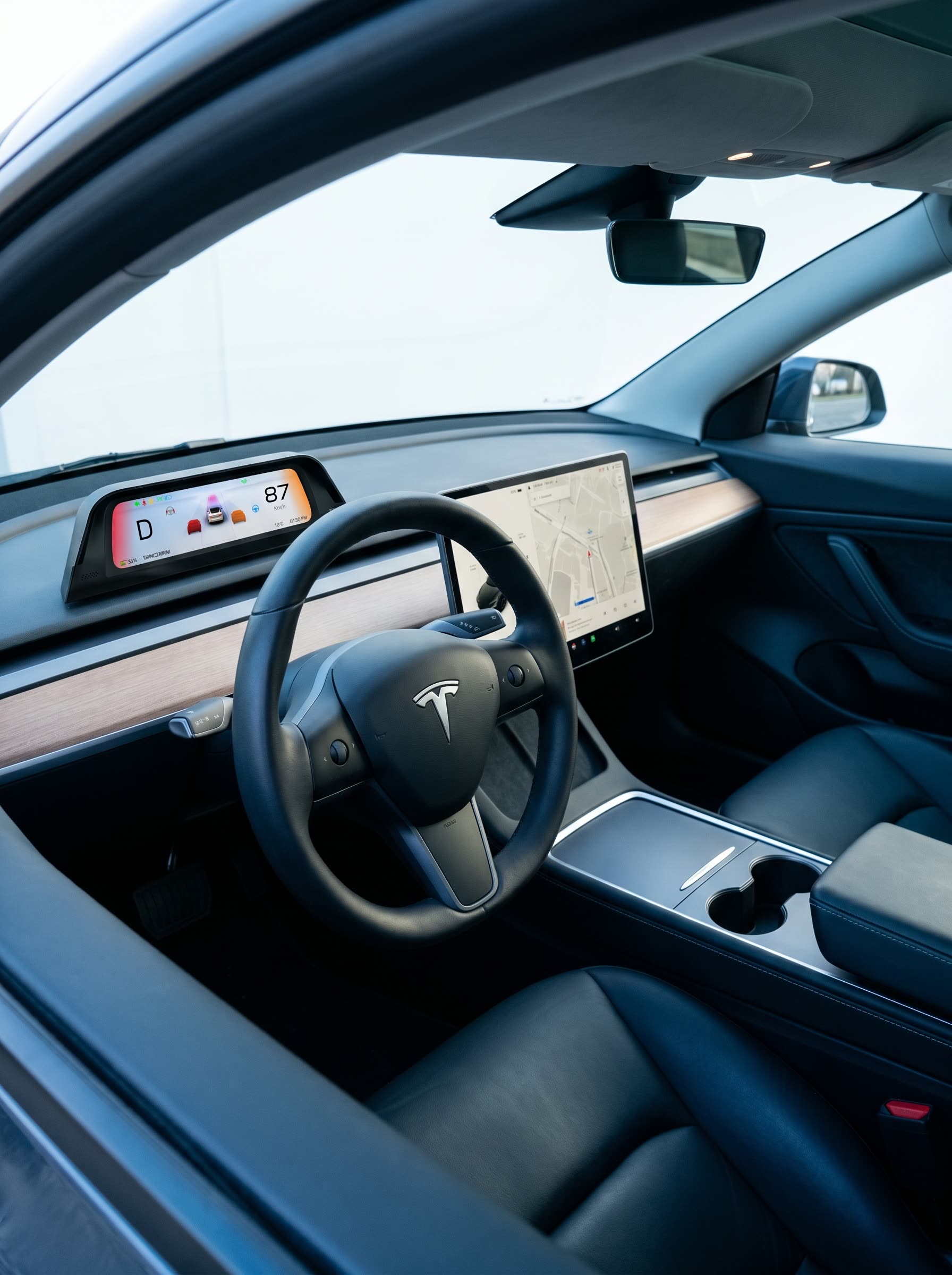 Innenraum eines linksgelenkten Tesla mit komplett sichtbarem Lenkrad und dem Display oben auf dem Armaturenbrett montiert, Gang D und Tempo 87 km/h.